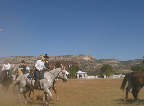 A group of horseback riders in western attire on a grassy field with mountains in the background under a clear blue sky.