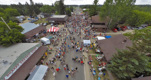 Aerial view of a bustling street fair with crowds, tents, and vendors lining the road amidst trees and buildings.