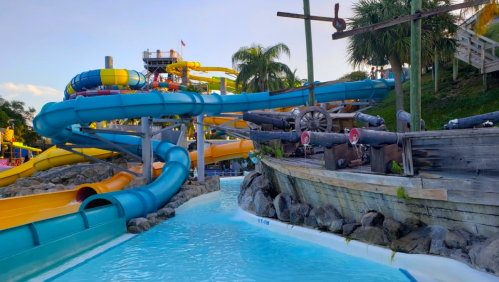 Colorful water slides twist above a lazy river, with a pirate ship and palm trees in the background.