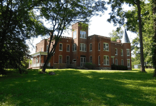 A large, brick castle-like house surrounded by trees and a grassy lawn on a sunny day.