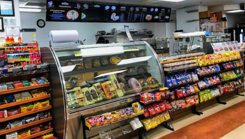 A deli display case filled with meats and cheeses, surrounded by shelves of snacks and drinks in a bright store.