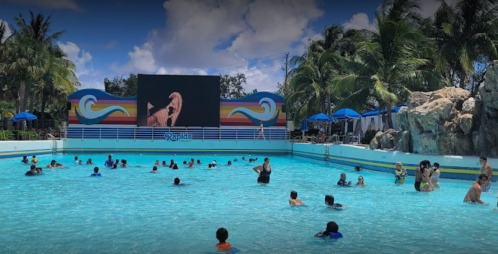 A vibrant water park scene with people swimming in a pool, a large screen, and palm trees under a sunny sky.