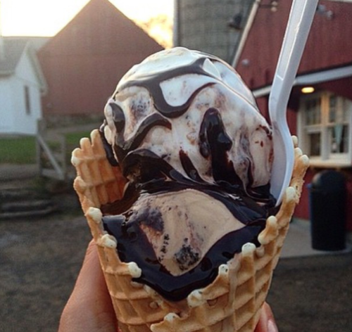 A hand holding a waffle cone filled with swirled chocolate and vanilla ice cream, drizzled with chocolate sauce.