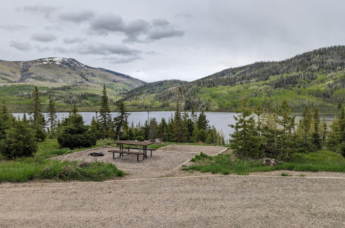 A serene lakeside view with picnic tables, surrounded by lush green trees and mountains under a cloudy sky.