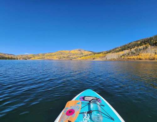 A paddleboard on calm water, surrounded by autumn-colored trees and mountains under a clear blue sky.