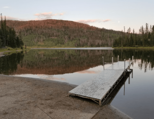 A wooden dock extends into a calm lake, surrounded by trees and a mountain, reflecting the serene landscape at dusk.