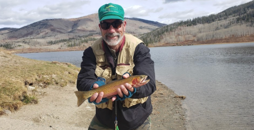 A smiling man in a cap holds a trout by a lake, surrounded by mountains and a cloudy sky.