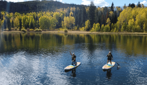 Two people paddleboarding on a calm lake surrounded by vibrant autumn trees and a clear blue sky.