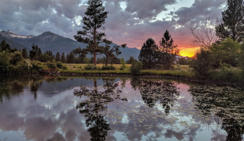 Sunset over a tranquil pond, with mountains and trees reflected in the water under a cloudy sky.
