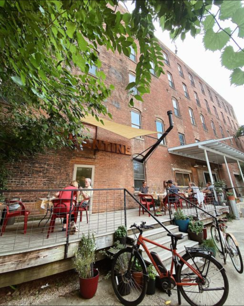 A cozy outdoor café with red chairs, a bike parked nearby, and greenery against a brick building backdrop.