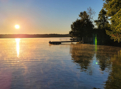Sunrise over a calm lake, with mist rising and a dock visible among trees on the shore.