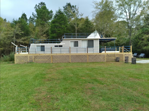 A mobile home with a deck surrounded by trees and a grassy area, featuring a lattice fence and outdoor seating.