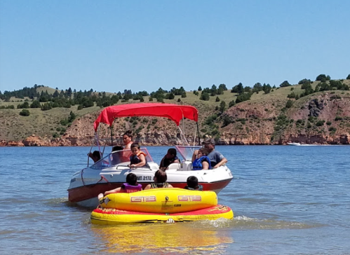 A boat with people on board pulls a yellow inflatable tube on a sunny day by a scenic lakeside.
