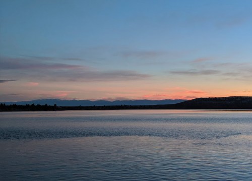 A serene lake at sunset, reflecting soft pink and blue hues with distant mountains on the horizon.