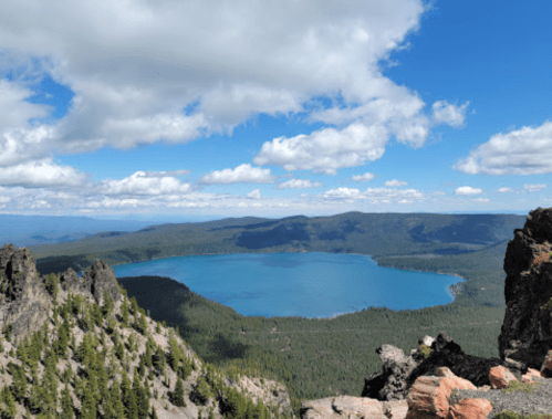A panoramic view of a blue lake surrounded by green forests and rocky terrain under a partly cloudy sky.