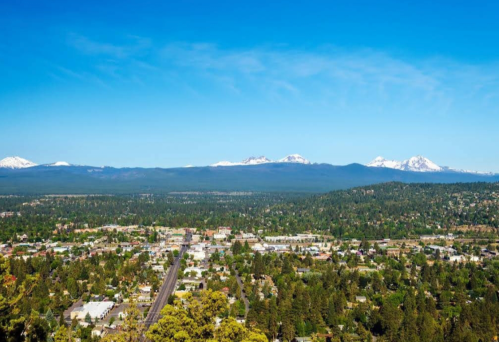 A panoramic view of a town surrounded by lush greenery and mountains under a clear blue sky. Snow-capped peaks in the distance.