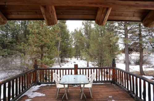 A wooden deck with a glass table and chairs, surrounded by snow-covered trees in a serene winter landscape.