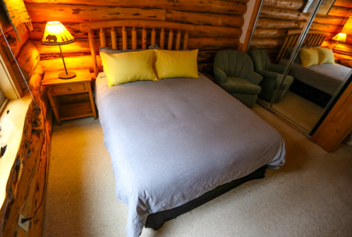 Cozy bedroom with a log cabin aesthetic, featuring a bed with yellow pillows, a green chair, and warm lighting.