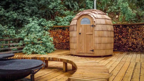 A wooden sauna hut with a rounded roof, surrounded by greenery and a wooden deck with a circular bench.