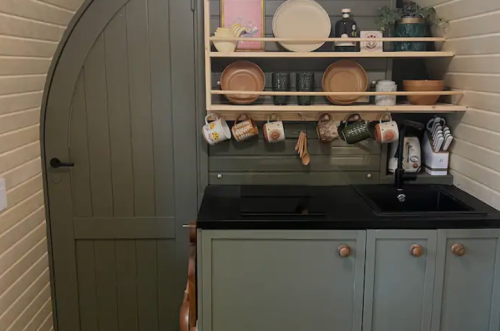 A cozy kitchen corner featuring a dark countertop, green cabinets, and shelves with dishes and mugs, beside an arched door.