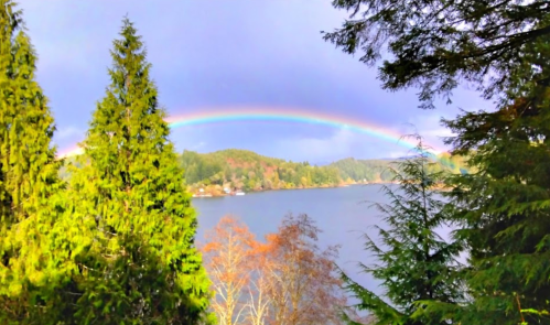 A vibrant rainbow arcs over a serene lake surrounded by lush green trees and autumn foliage.