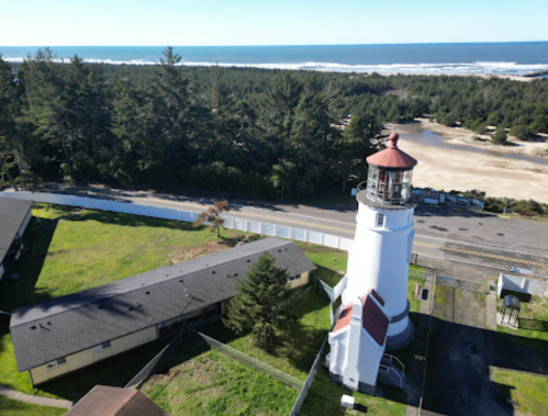 Aerial view of a lighthouse near the coast, surrounded by trees and a sandy beach under a clear blue sky.
