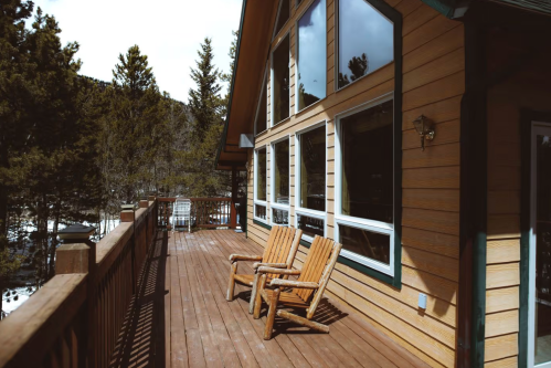 A wooden deck with two chairs overlooks a forested area, featuring large windows and a cozy cabin atmosphere.