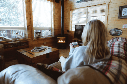 A cozy cabin interior with a person holding a mug, sitting on a couch near a warm fireplace and large windows.
