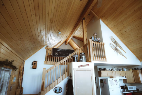 A woman stands on a balcony in a wooden cabin with high ceilings and rustic decor, overlooking the kitchen below.