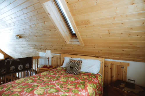 Cozy attic bedroom with a wooden ceiling, floral bedding, and a skylight, featuring a lamp and decorative pillows.