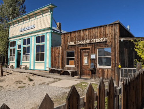 Two historic buildings side by side: a colorful shop and a rustic wooden store, set against a clear blue sky.