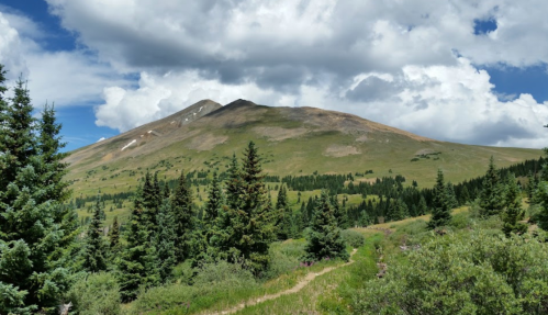 A scenic mountain landscape with green trees in the foreground and a cloudy blue sky above.