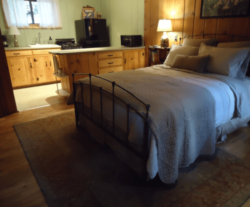 Cozy bedroom with a metal bed frame, soft bedding, and wooden walls, featuring a kitchenette in the background.