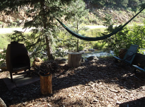 A serene outdoor scene with a hammock, fire pit, and chair by a stream, surrounded by trees and rocky terrain.
