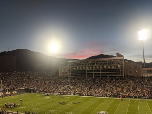 A football stadium filled with fans at dusk, with mountains in the background and bright stadium lights illuminating the field.