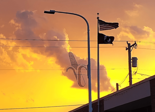 A sunset sky with orange clouds, featuring an American flag and a POW/MIA flag on poles. A banana-shaped sculpture is visible.