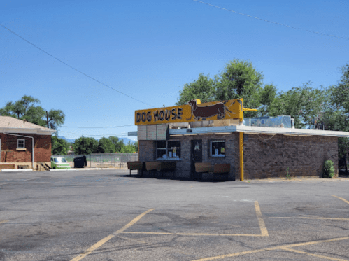 A small, brick building with a bright yellow sign reading "Dog House," set in a parking lot under a clear blue sky.