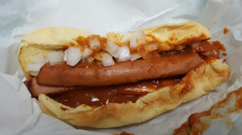 A hot dog in a bun topped with chili and diced onions, served in white paper.