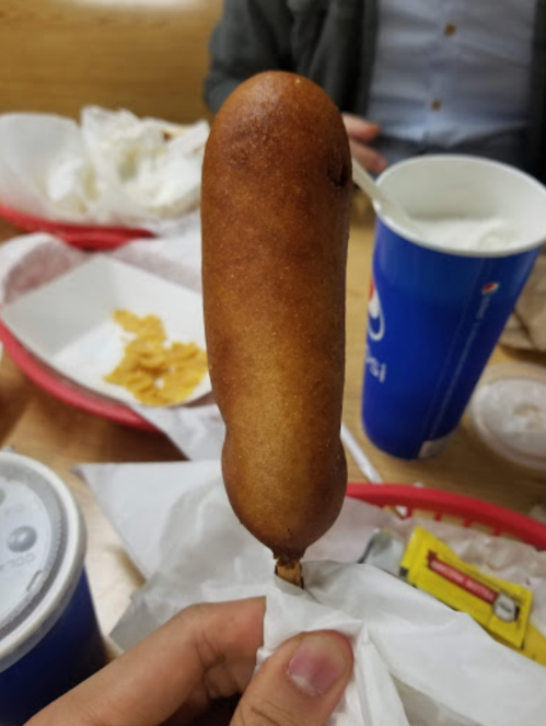 A hand holding a golden-brown corn dog on a stick, with food wrappers and drinks in the background.
