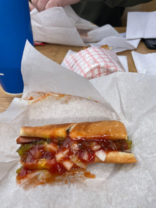 A hot dog in a bun topped with ketchup, mustard, and diced onions, resting on white paper with a blue drink nearby.