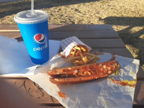 A hot dog with sauce, a small serving of fries in paper, and a Pepsi drink on a wooden table outdoors.