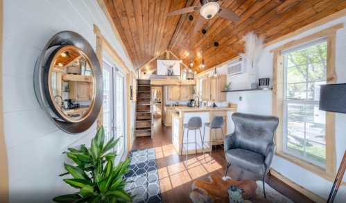 Cozy interior of a tiny home featuring wooden beams, a staircase, kitchen, and stylish decor with plants.