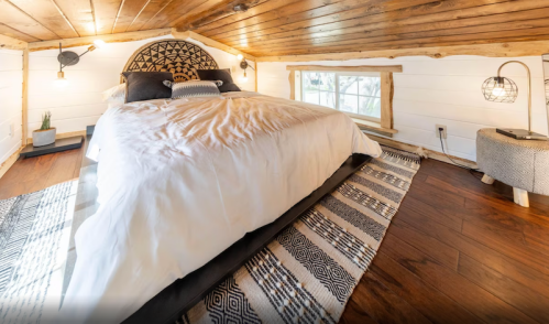Cozy attic bedroom with a large bed, wooden ceiling, and decorative rug, featuring natural light from a window.