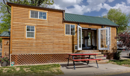 A cozy wooden tiny house with a green metal roof, featuring open doors and a picnic table outside.