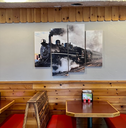 A triptych of a vintage steam train hangs on a wooden wall in a cozy diner with red booths and tables.