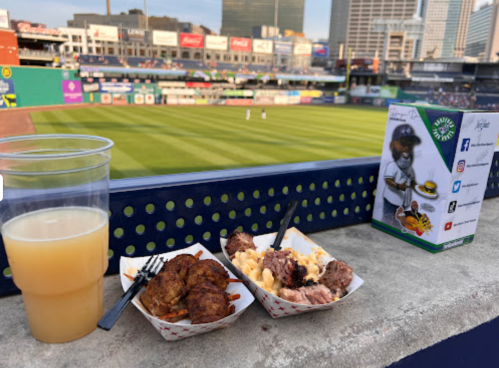 A view of a baseball field with food and a drink on a ledge, featuring meatballs, macaroni, and a beer.