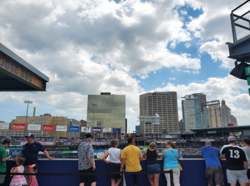 A crowd watches a baseball game against a backdrop of city buildings and a partly cloudy sky.