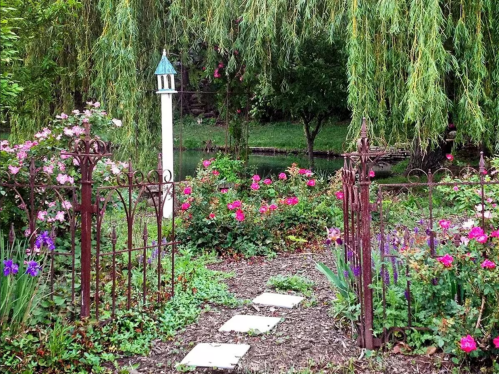A charming garden path with colorful flowers, leading through an ornate iron gate to a serene pond.