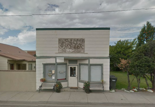 A small, two-story building with a sign reading "Self Surrender," surrounded by greenery and a clear sky.