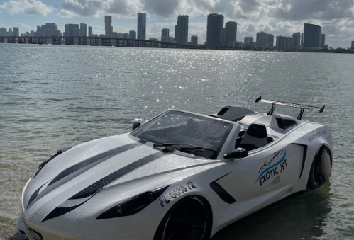 A sleek white sports car parked by the water with a city skyline in the background under a cloudy sky.
